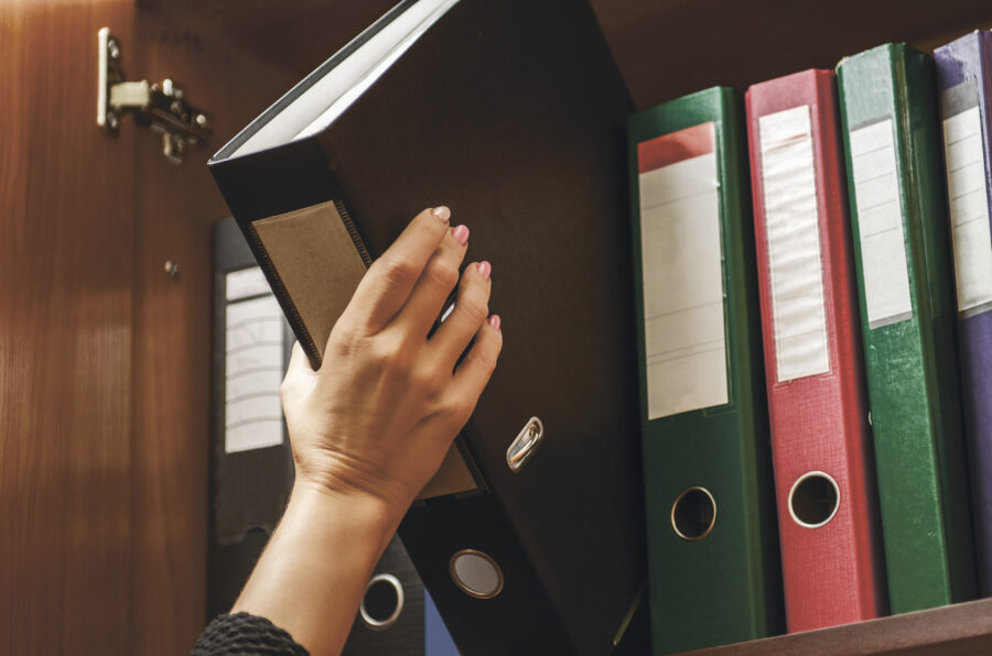 a woman officer pick a binder of document on the row of file folders and paper that nicely management system on the office's shelves and holing it with her hand