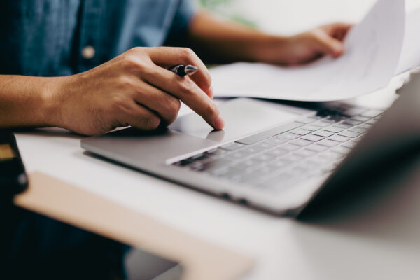 While reading a business document, a businessman's hand is on the trackpad, typing on a laptop keyboard. A corporate man, a lawyer, works in a modern office. Business and technology concepts