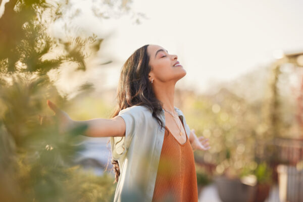 Young woman with arms outstretched breathing in fresh air during sunrise at the balcony. Healthy girl enjoying nature while meditating during morning with open arms and closed eyes. Mindful woman enjoying morning ritual while relaxing in outdoor park.