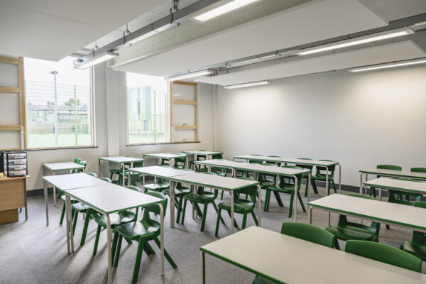 Wide angle view of unoccupied educational environment with desks accommodating two students each, chairs, windows, and overhead LED lighting.